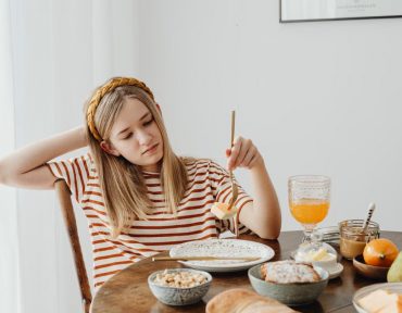 Teen girl in striped shirt at breakfast table with fruits, bread, and juice. Casual morning setting.